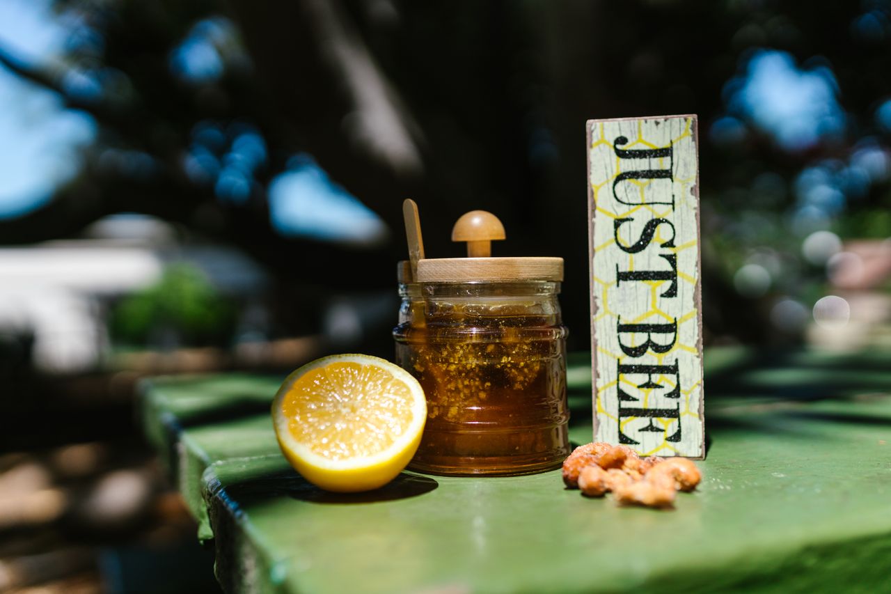 Close-up of a honey jar, lemon, and 'Just Bee' sign on a green table outdoors.